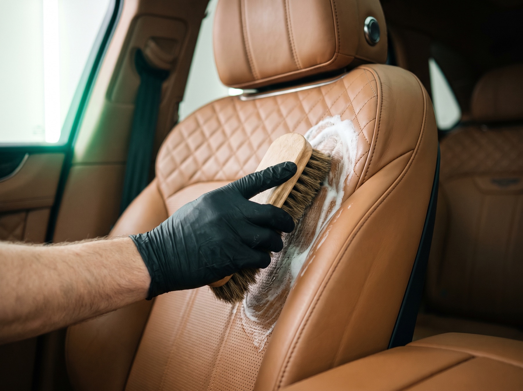 Gloved hand cleaning a tan leather car seat with a soft brush and foam